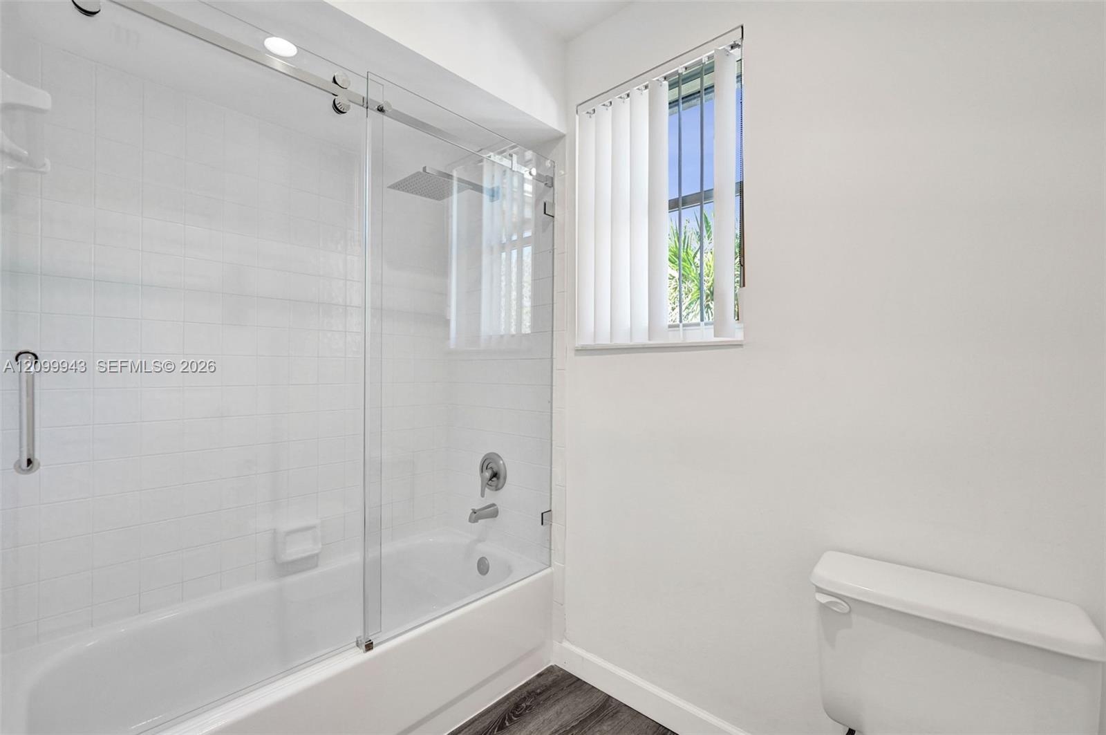 Bathroom featuring a tub-shower combination with frameless glass sliding doors, polished chrome fixtures, square wall tiling, and wood-finish flooring