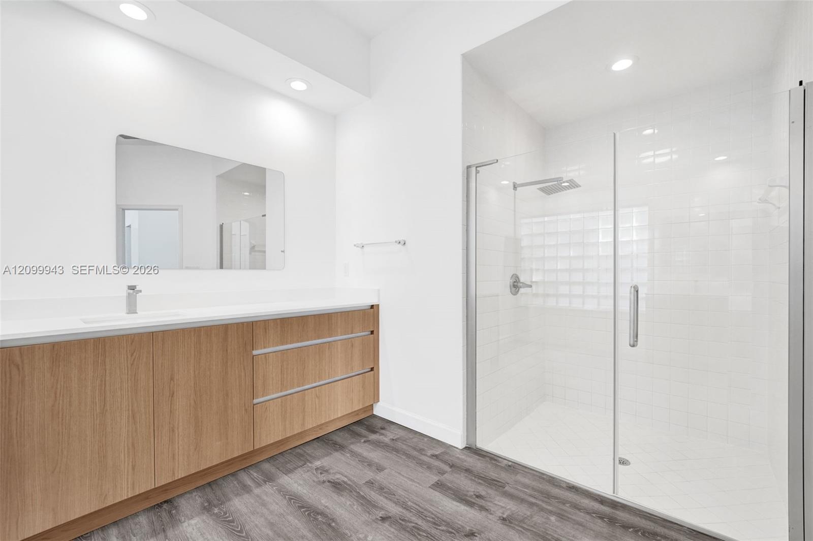 Floating wood-finish vanity with a white countertop and rectangular sink, frameless glass-enclosed shower, polished chrome fixtures, and wood-finish f
