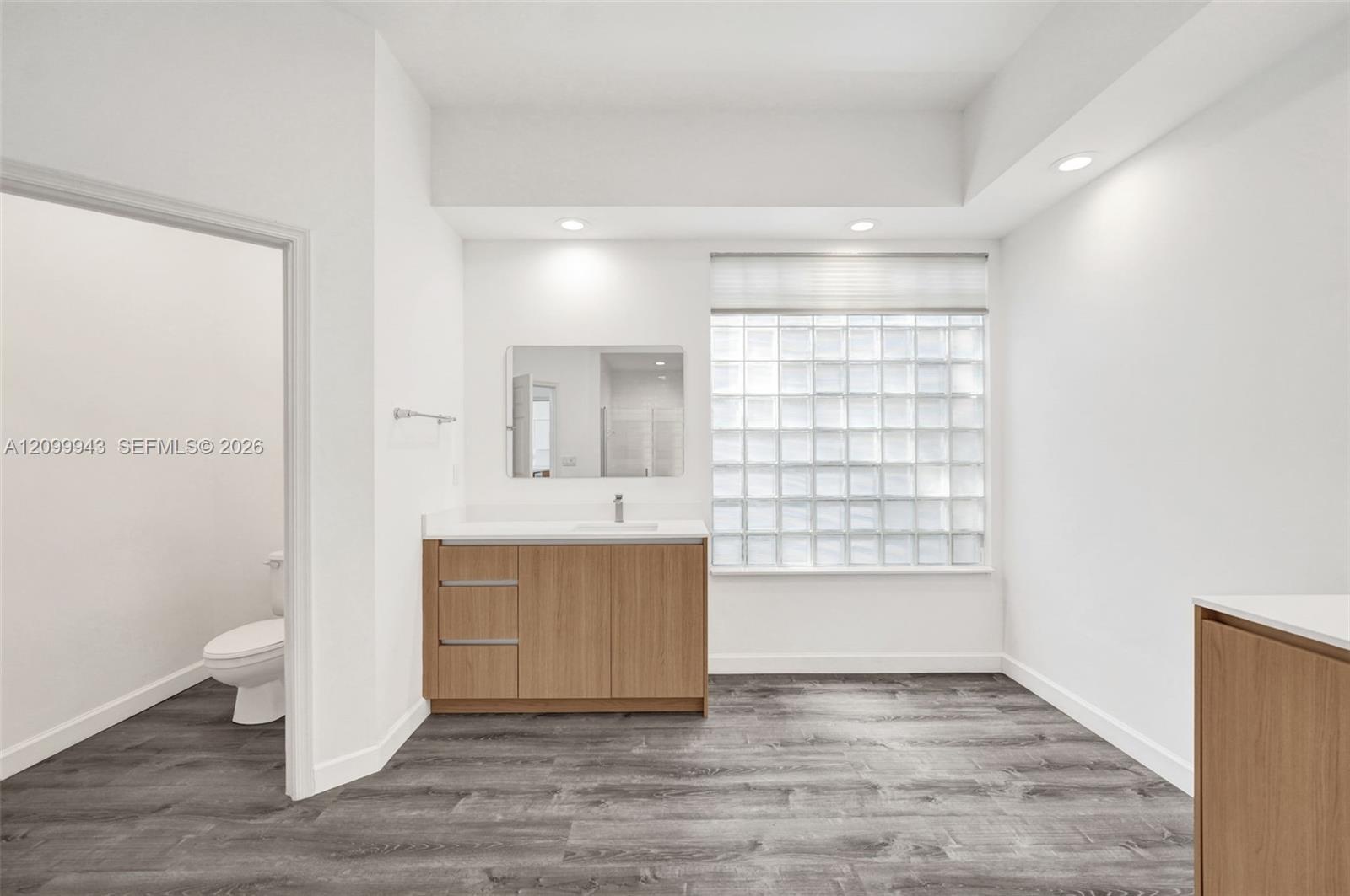 Wood-finish cabinetry with a white surface, rectangular wall-mounted mirror, glass block window, recessed lighting, and wood-finish flooring