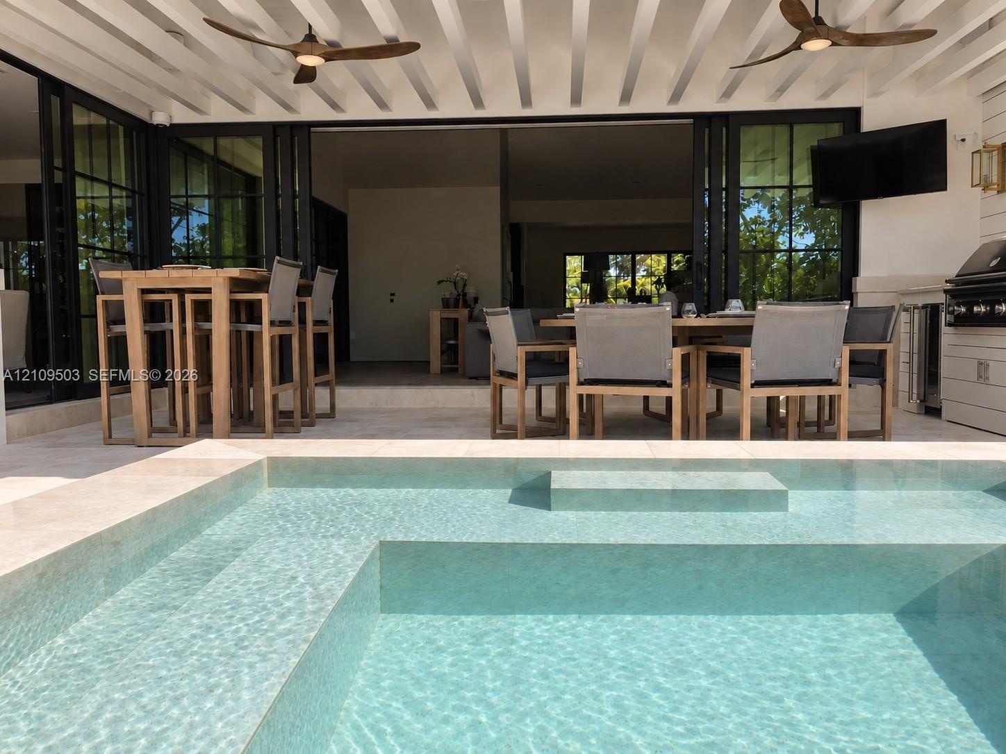 Covered patio featuring an integrated outdoor kitchen, multi-panel retractable glass doors, dual ceiling fans, and an inground swimming pool with ston