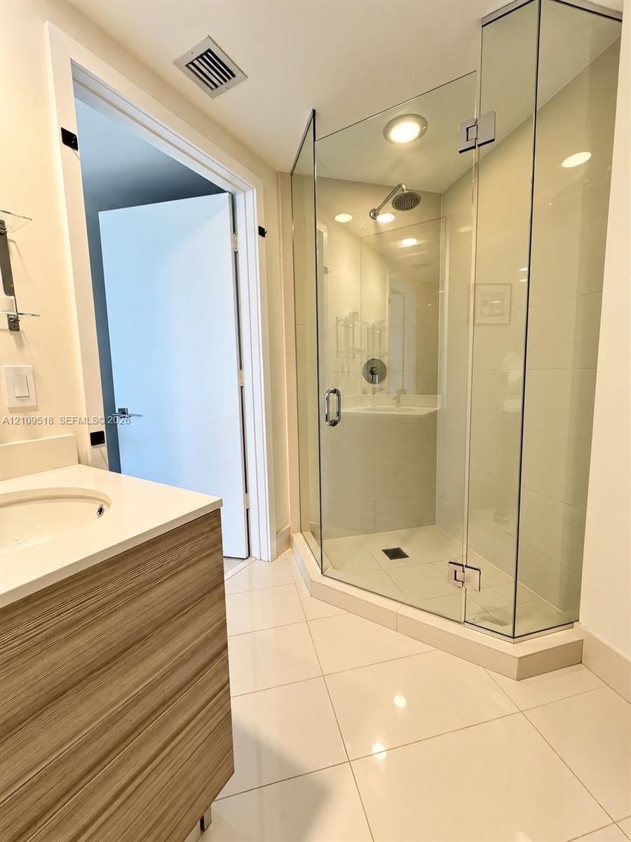 Glass-enclosed corner shower with a polished metal showerhead, vanity with wood-finish cabinetry and light-colored countertop, and large-format tile f