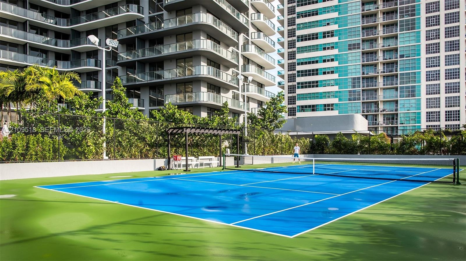 Tennis court with high-contrast dual-tone hard surfacing, perimeter chain-link fencing, structural pergola, and integrated lighting