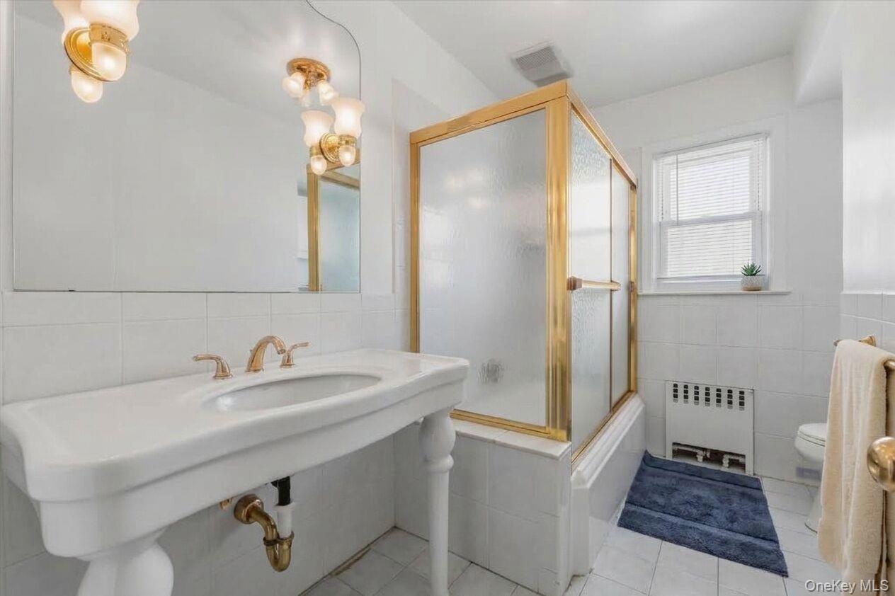 Pedestal sink with brass-finish faucet, gold-tone shower enclosure, white tile walls, and suspended lighting fixtures