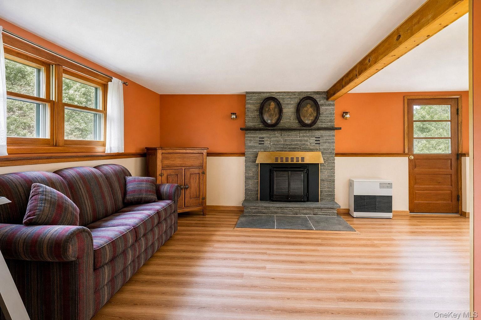 Living area featuring stone fireplace with wood stove insert and dual windows for view of front yard and pond, exposed ceiling beam.