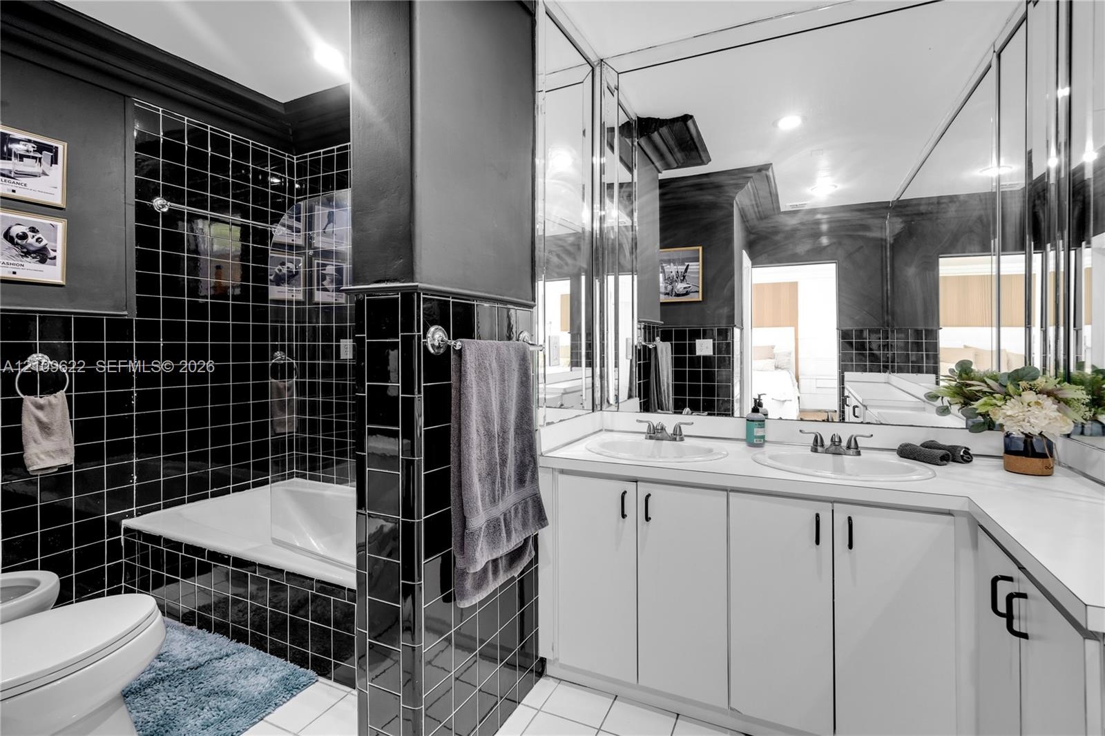 Bathroom featuring black square wall tiles, dual-sink vanity with white countertop, full-width mirror, and deep soaking tub with integrated shower