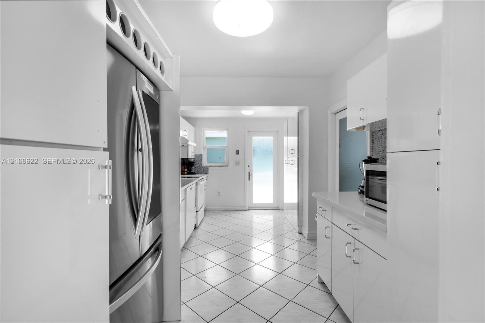 Kitchen featuring high-gloss white cabinetry, white stone-surface countertops, stainless steel refrigerator, tile flooring, and a glass-paneled exteri