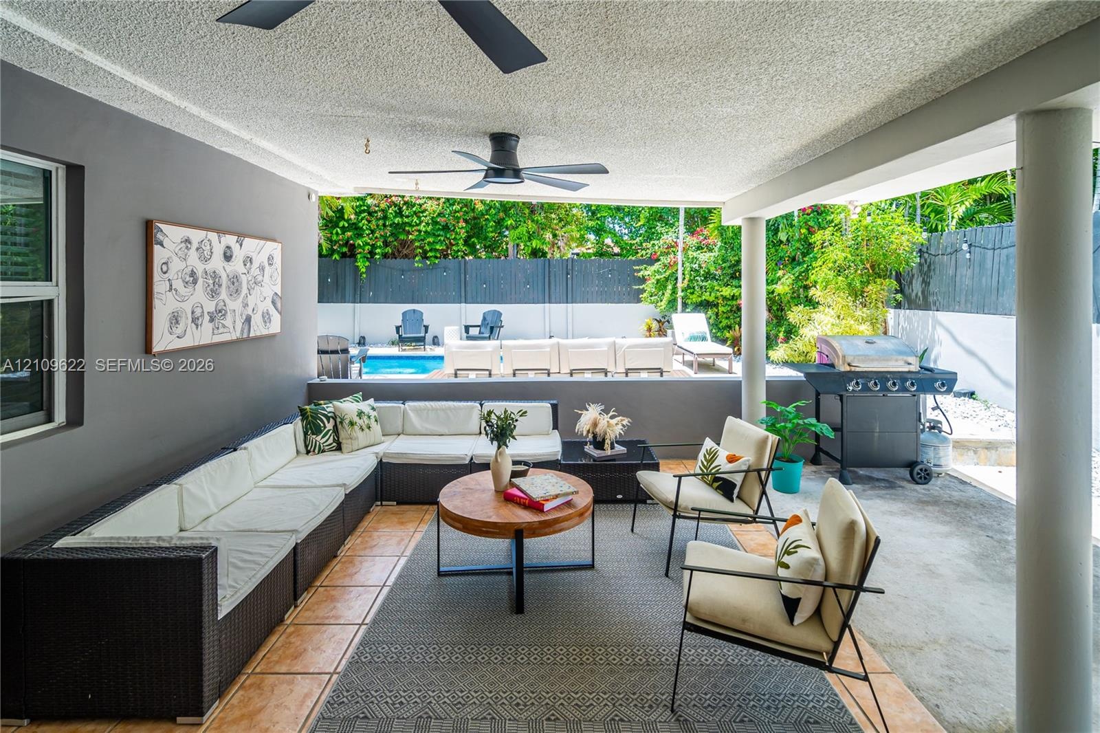 Covered patio featuring tiled flooring, two ceiling fans, and structural support columns, opening to a swimming pool and perimeter fencing
