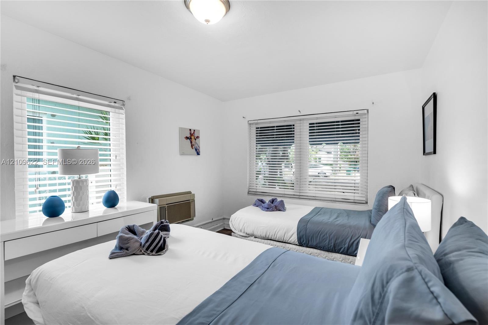 Bedroom featuring dual windows with horizontal blinds, neutral wall paint, flush-mount lighting, and an integrated through-wall air conditioning unit