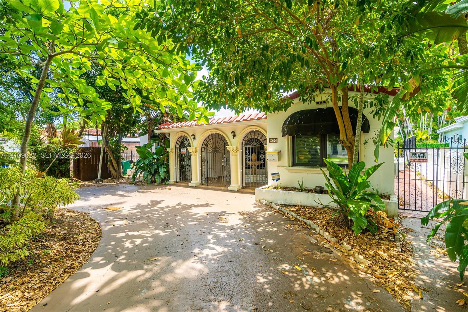 Exterior facade featuring arched entryways with decorative iron security grilles, a barrel-tile roofline, concrete driveway, and mature landscaping