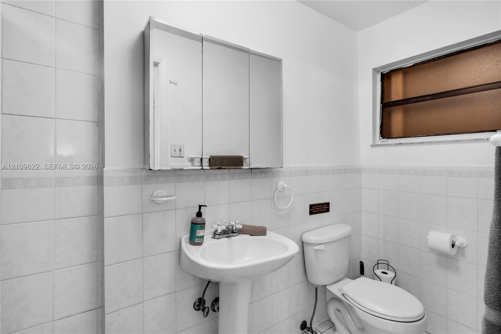 Bathroom featuring white tile wall cladding, wall-mounted three-panel mirrored medicine cabinet, porcelain pedestal sink, and fixed-pane window