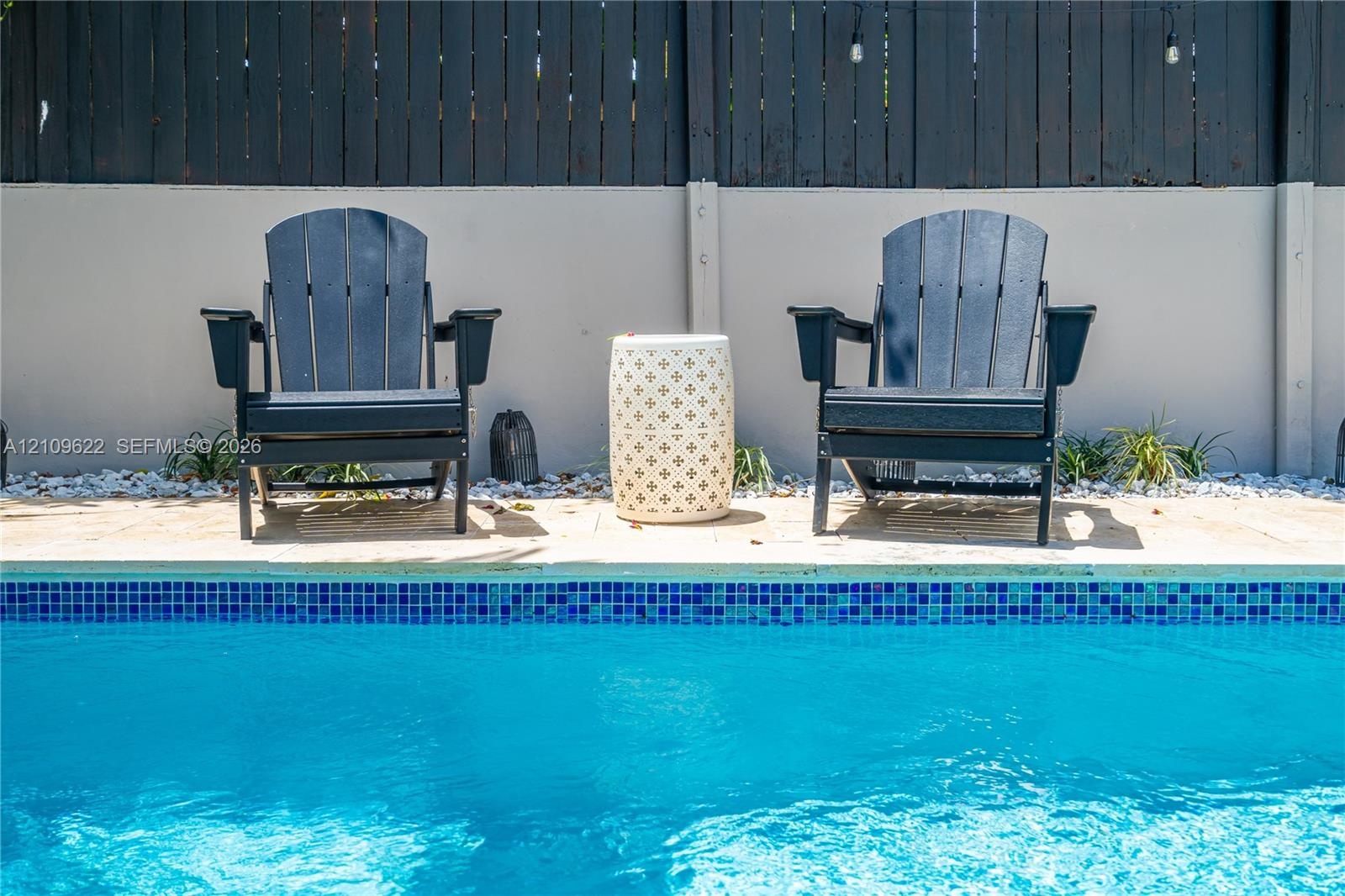 Swimming pool with blue glass tile waterline, stone-paved decking, light-toned masonry perimeter wall, and dark-finished vertical board fencing