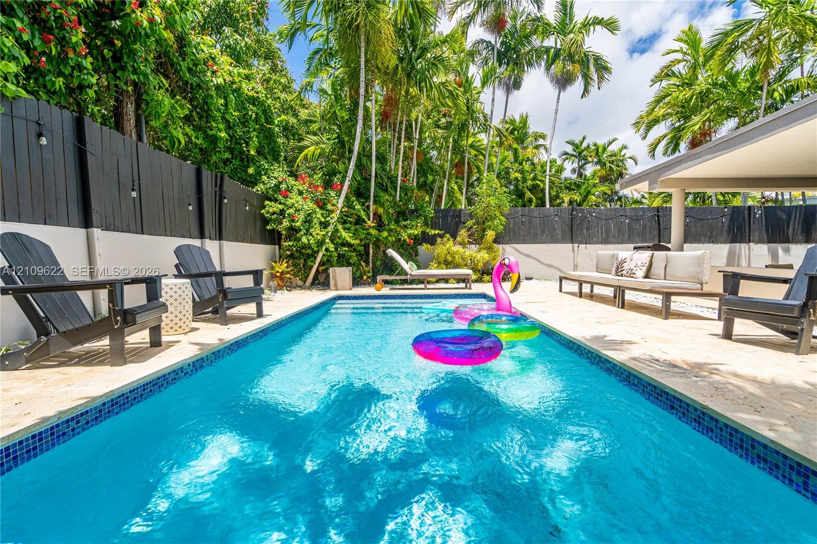 In-ground swimming pool with blue tile border, limestone-style paver decking, covered patio area, and privacy fencing complemented by mature palm tree