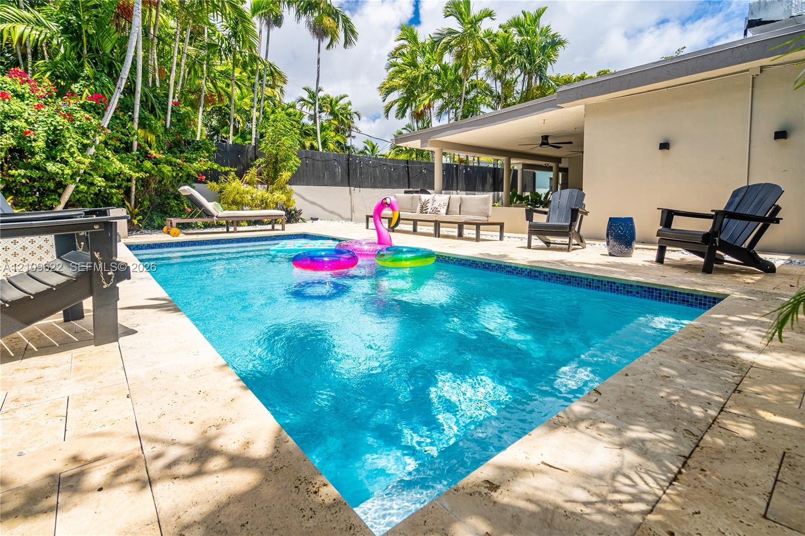 Swimming pool with stone paver decking and covered patio area featuring a ceiling fan, surrounded by privacy fencing and mature palm trees
