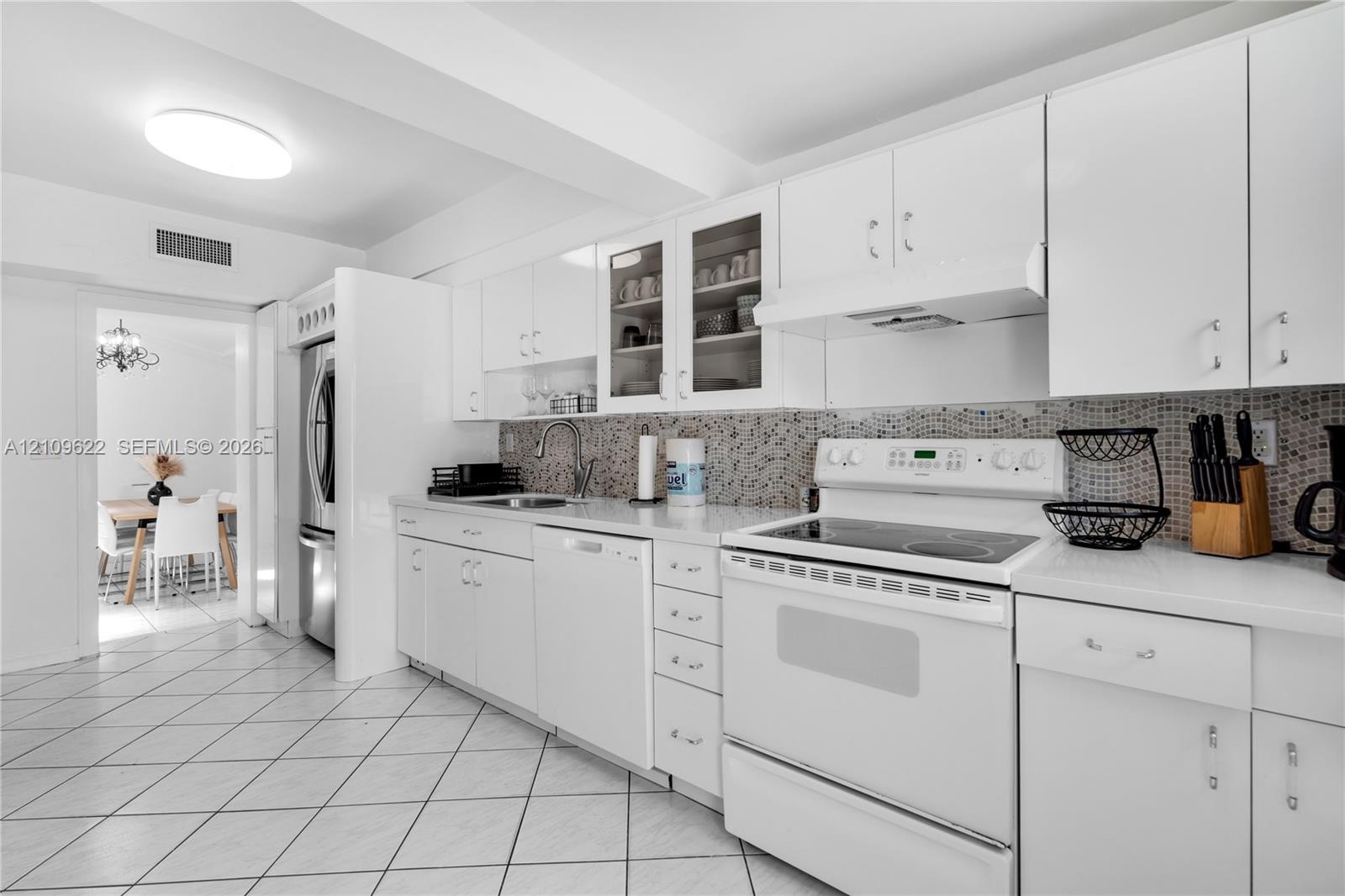 Kitchen featuring white cabinetry with glass-front display uppers, mosaic tile backsplash, solid-surface countertops, tile flooring, and integrated wh