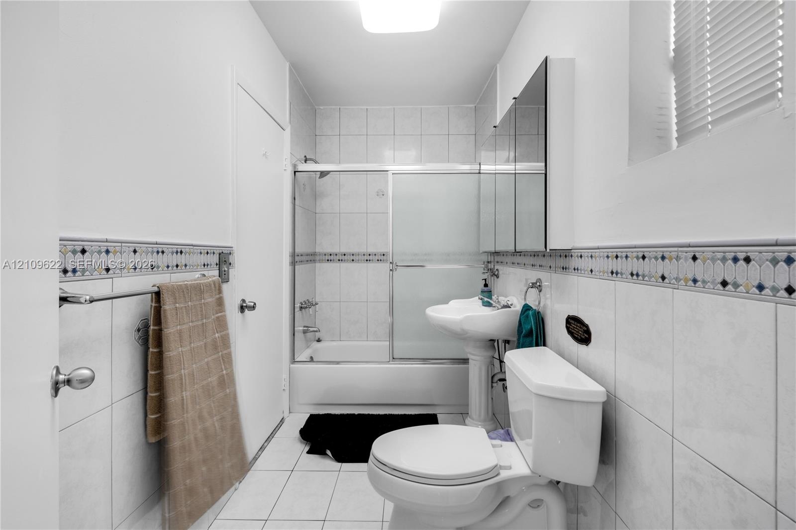 Bathroom featuring white tile flooring, pedestal sink, tub-shower combination with sliding glass doors, and decorative tile accent wall detailing