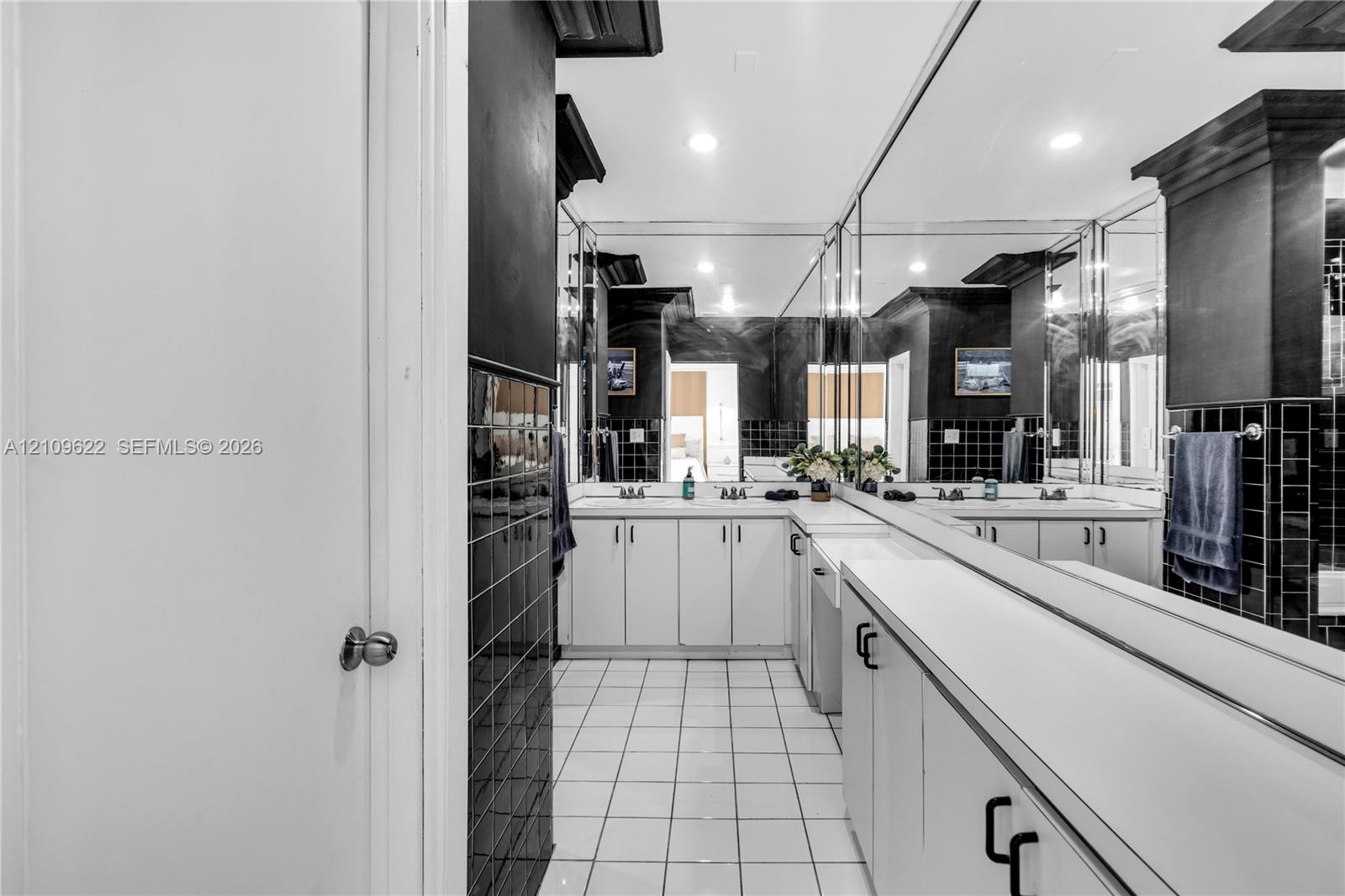 Dual-sink vanity with white cabinetry and solid surface countertops, expansive wall-to-wall mirror, and contrasting black tile backsplash and wall acc