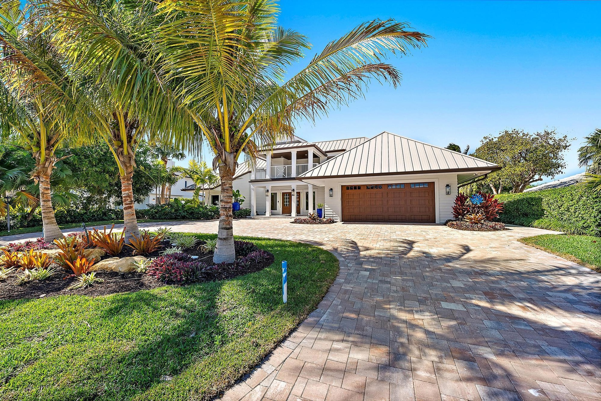 Exterior featuring a standing-seam metal roof, paver driveway, three-car garage, covered entry with a second-story balcony, and professional landscapi