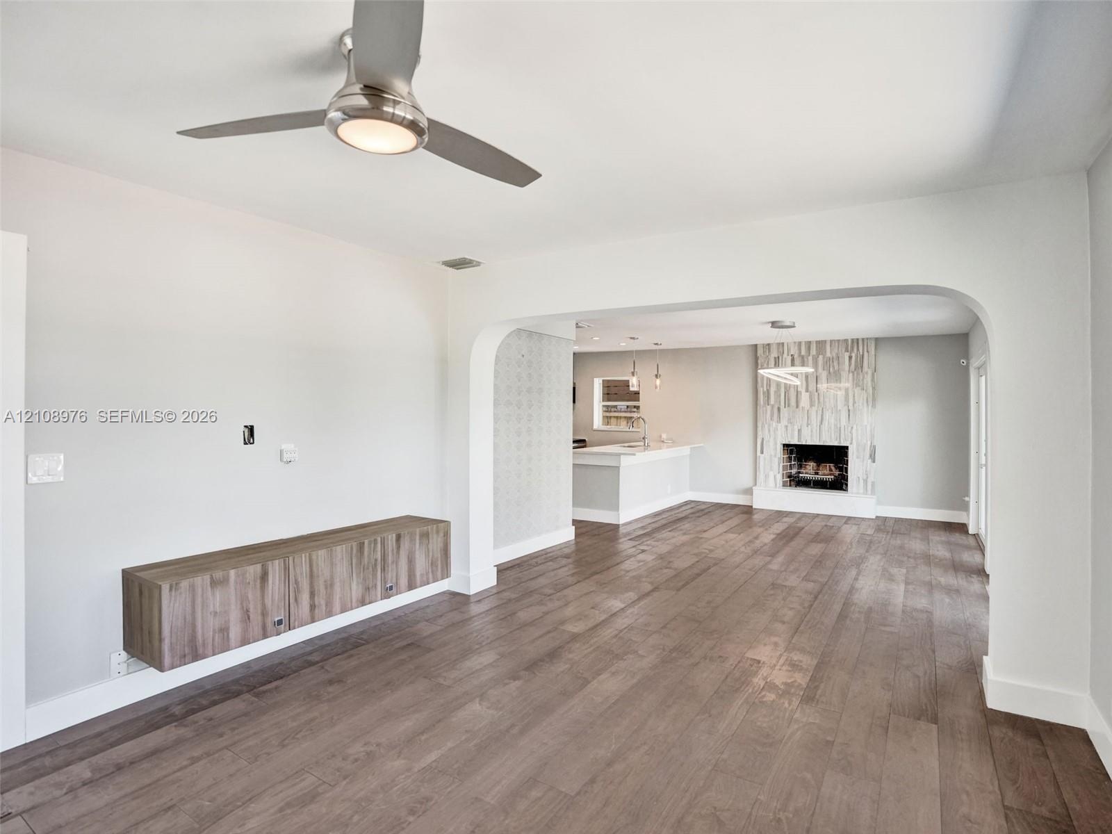 Wood-finish flooring, brushed-nickel ceiling fan with integrated lighting, wall-mounted wood-finish cabinetry, arched entryway, and fireplace with ver