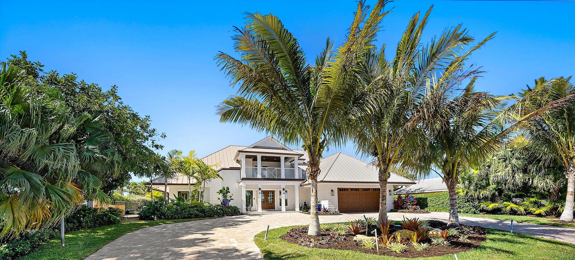 Two-story exterior featuring a metal roof, balcony with decorative railings, multi-car garage, and a paved circular driveway surrounded by mature palm