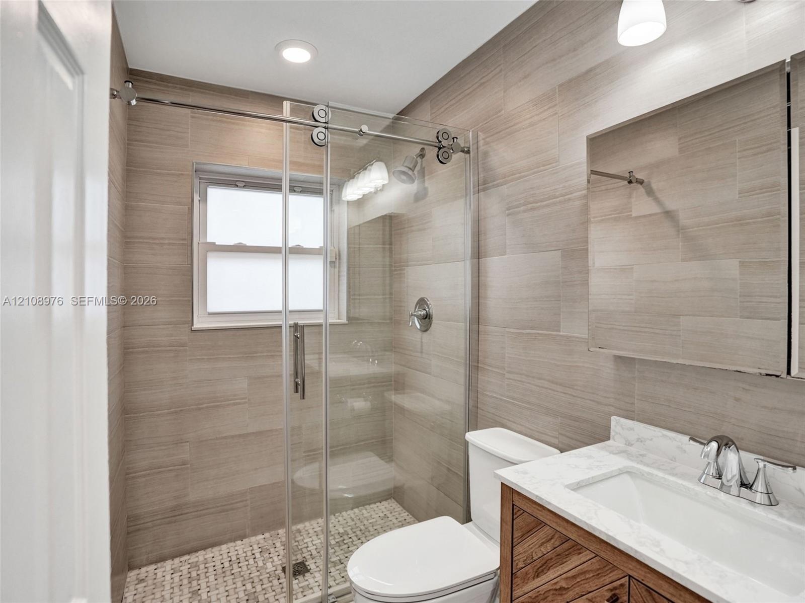 Walk-in shower with sliding glass door, mosaic floor tiling, full-height wall tile, wood-finish vanity with stone countertop, and recessed ceiling lig