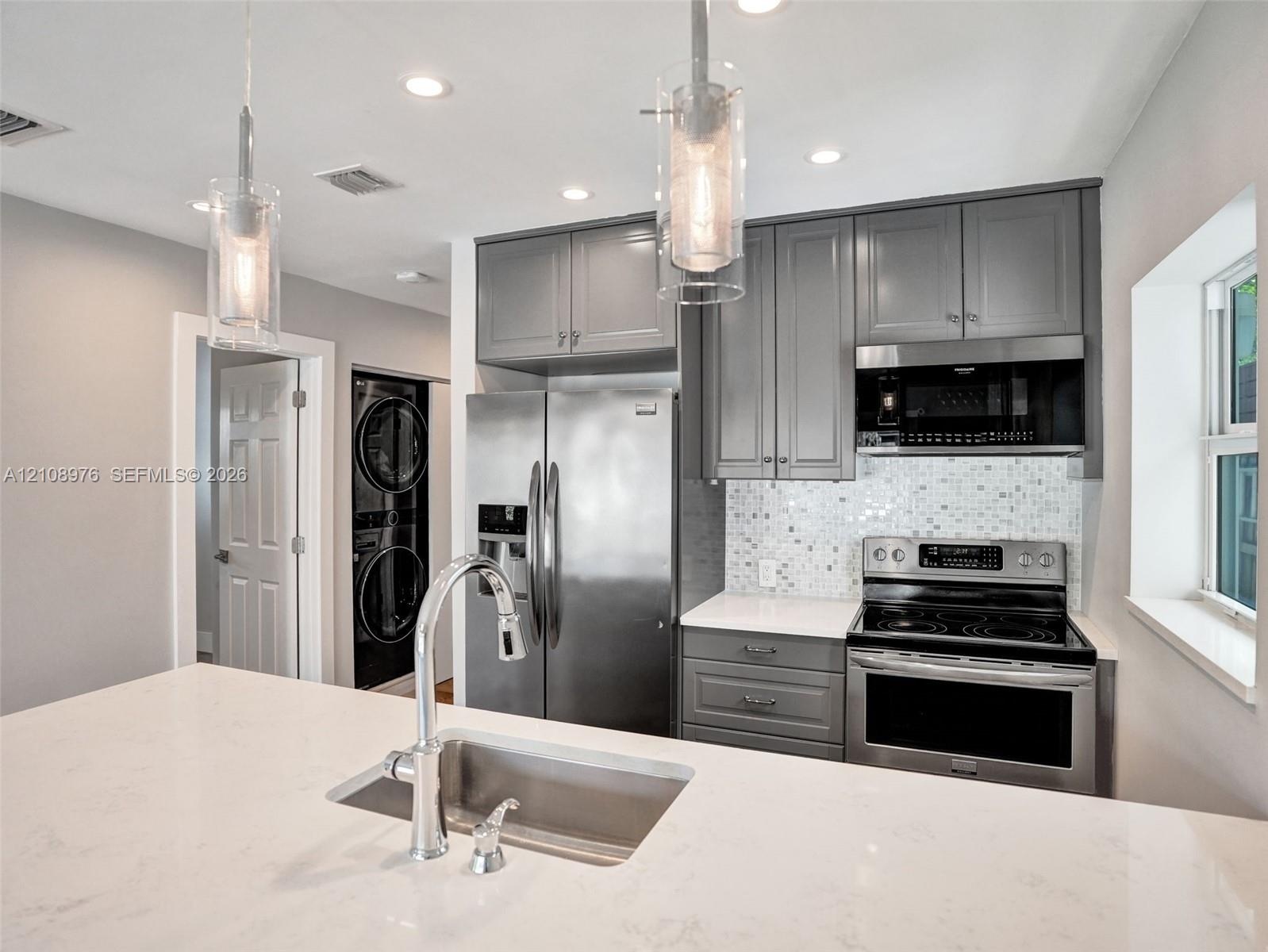 Kitchen featuring grey shaker-style cabinetry, white stone-finish countertops, stainless steel appliances, mosaic tile backsplash, and glass pendant l