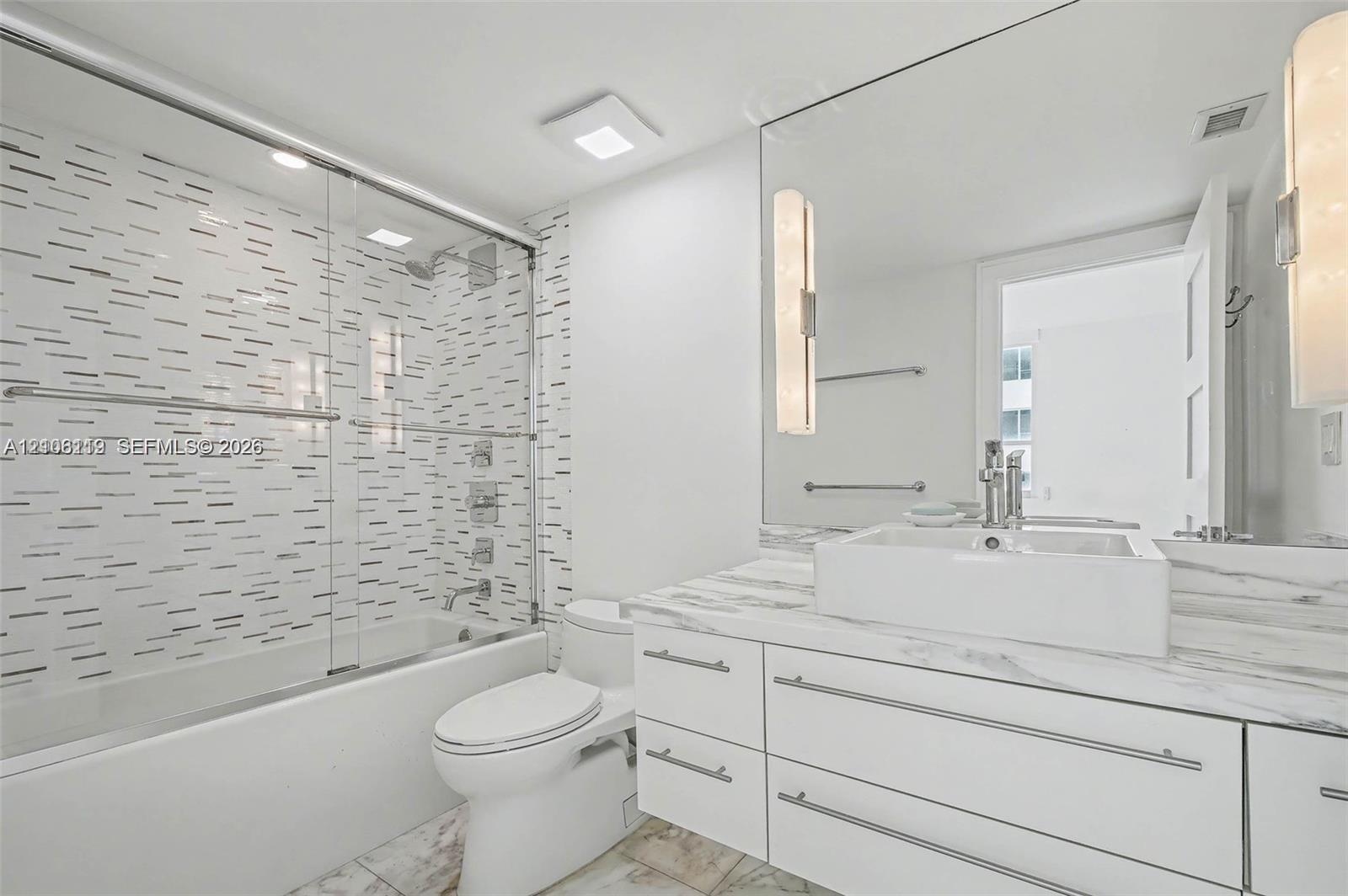 Bathroom featuring a white vanity with a stone-finish countertop and square vessel sink, full-width wall mirror with integrated lighting, glass-enclos