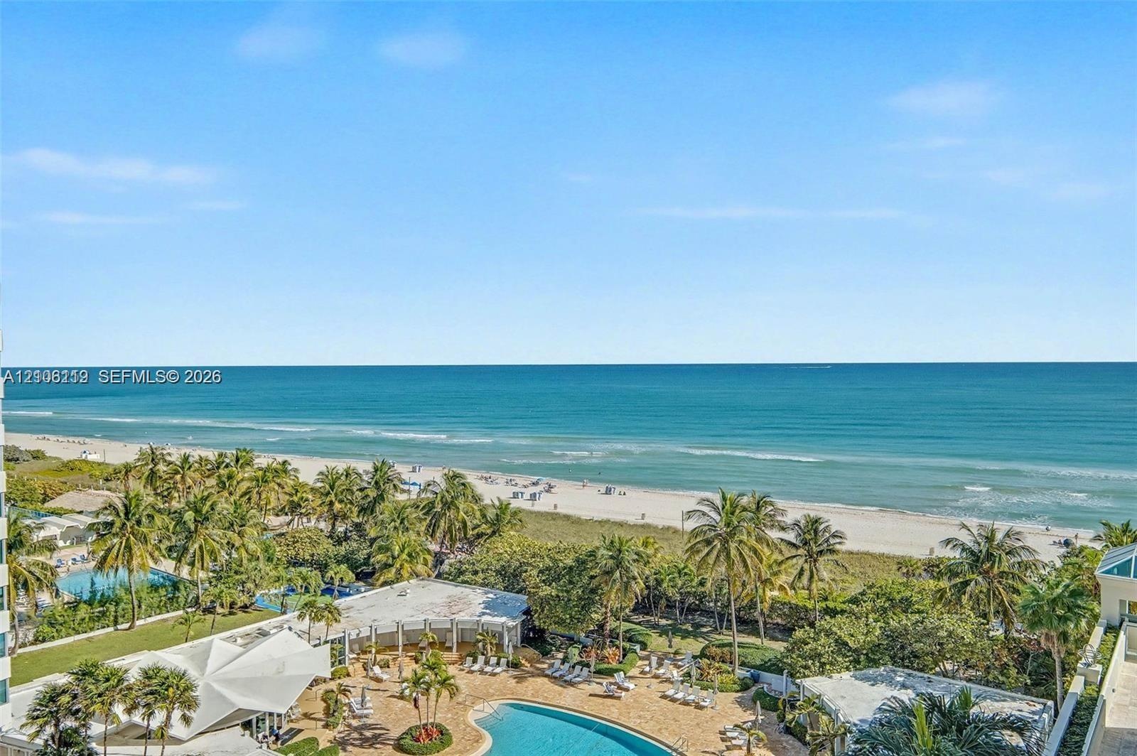 Elevated perspective featuring direct oceanfront access, sandy beach frontage, mature palm trees, and an expansive outdoor swimming pool with surround