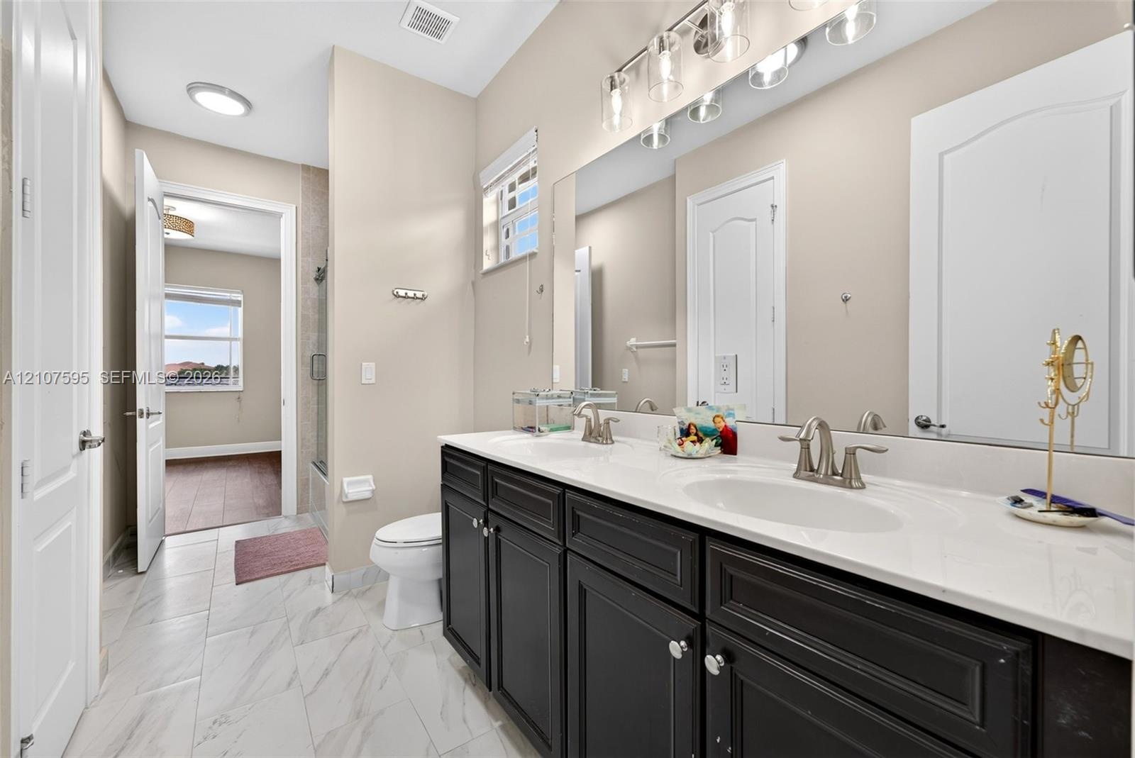 Double vanity with dark cabinetry, stone-finish countertops, dual under-mount sinks, and large wall-mounted mirror, featuring porcelain tile flooring 