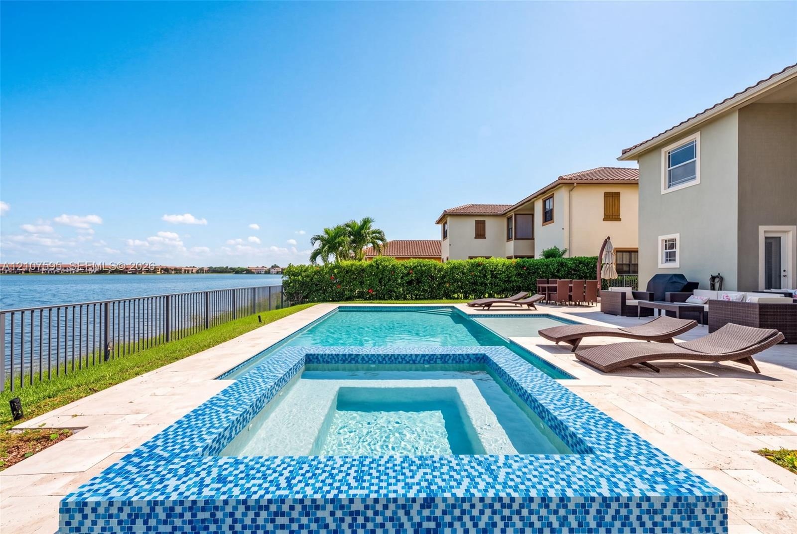 In-ground swimming pool and spa featuring blue tile accents, surrounded by a light-toned stone paver deck and metal perimeter fencing