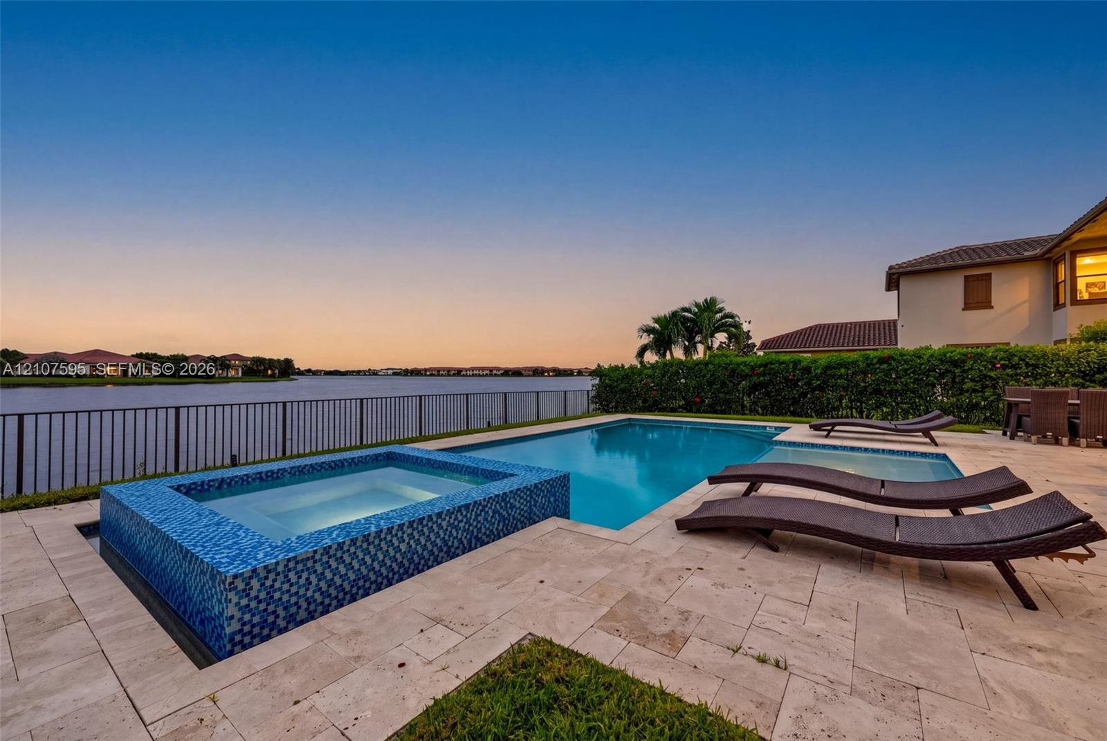 In-ground swimming pool and raised spa with blue tile accents, surrounded by a stone-paver deck, enclosed by a perimeter fence, overlooking a water fe