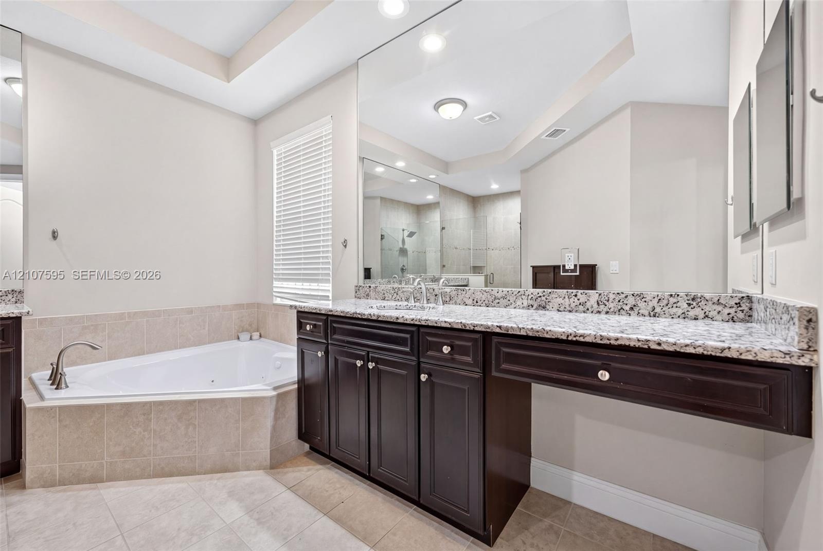 Built-in dark wood vanity featuring a speckled stone countertop, large-scale wall-mounted mirror, corner jetted soaking tub with tile surround, and ne