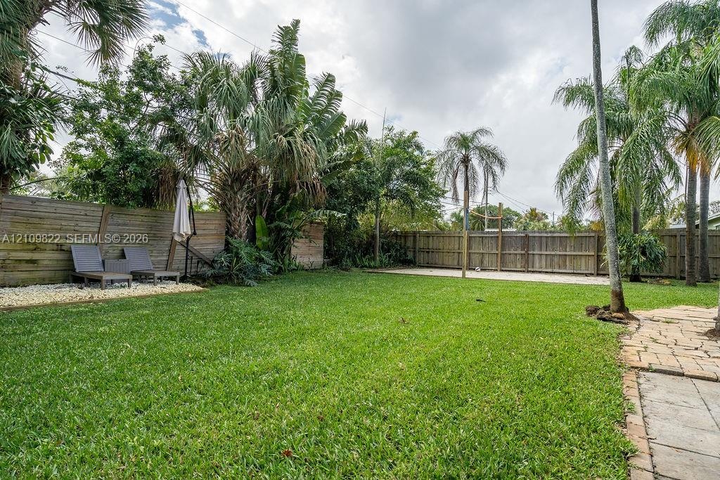 Expansive backyard featuring a lush lawn, mature palm trees, perimeter wood fencing, stone paver accents, and decorative pebble ground cover