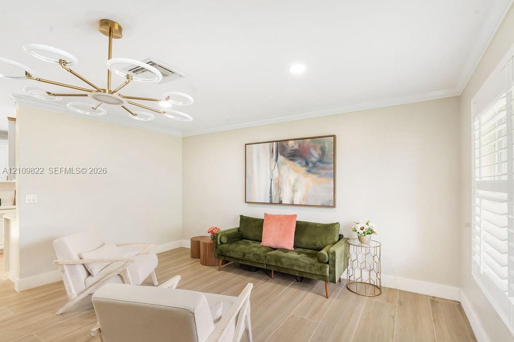 Spacious room featuring wood-finish tile flooring, crown molding, tiered gold-tone chandelier with integrated circular lighting, and plantation shutte