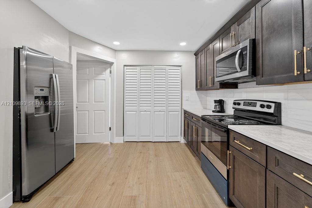 Kitchen featuring wood-finish flooring, dark-toned cabinetry with brass-finish hardware, stainless steel appliances, light-toned countertops, and a wh