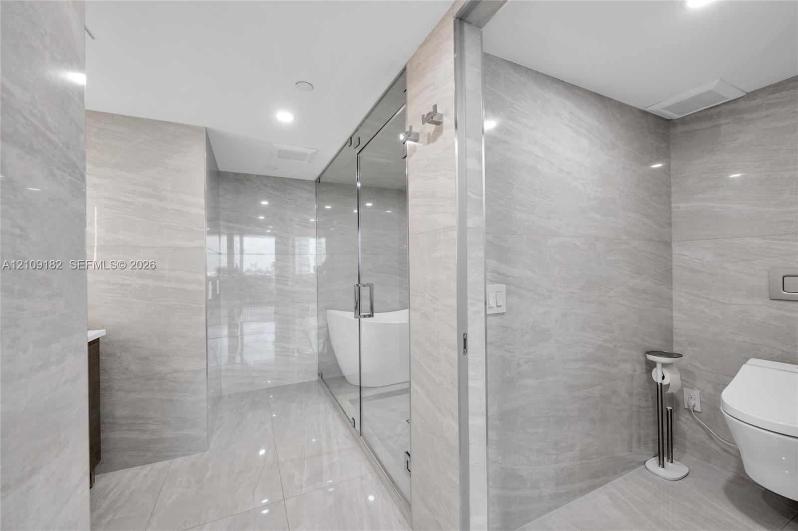 Dual-sink vanity with a solid-surface countertop, dark cabinetry, recessed lighting, wall-mounted backlit mirrors, and floor-to-ceiling stone-tiled sh