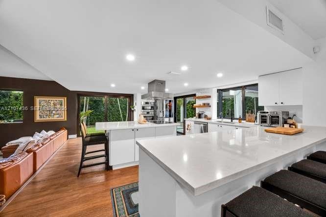 Modern kitchen featuring an expansive central island with quartz countertops, custom white cabinetry, stainless steel range hood, recessed lighting, a
