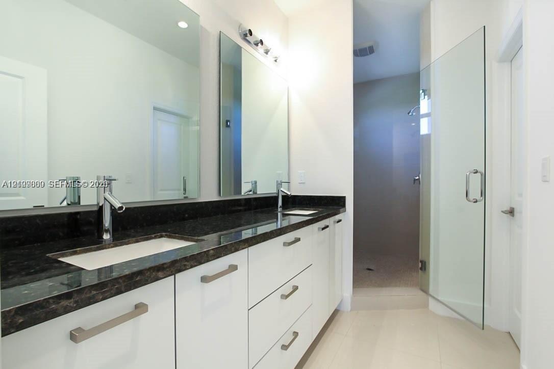 Dual-sink vanity featuring stone countertops, white cabinetry with horizontal hardware, two wall-mounted mirrors, and a glass-enclosed shower