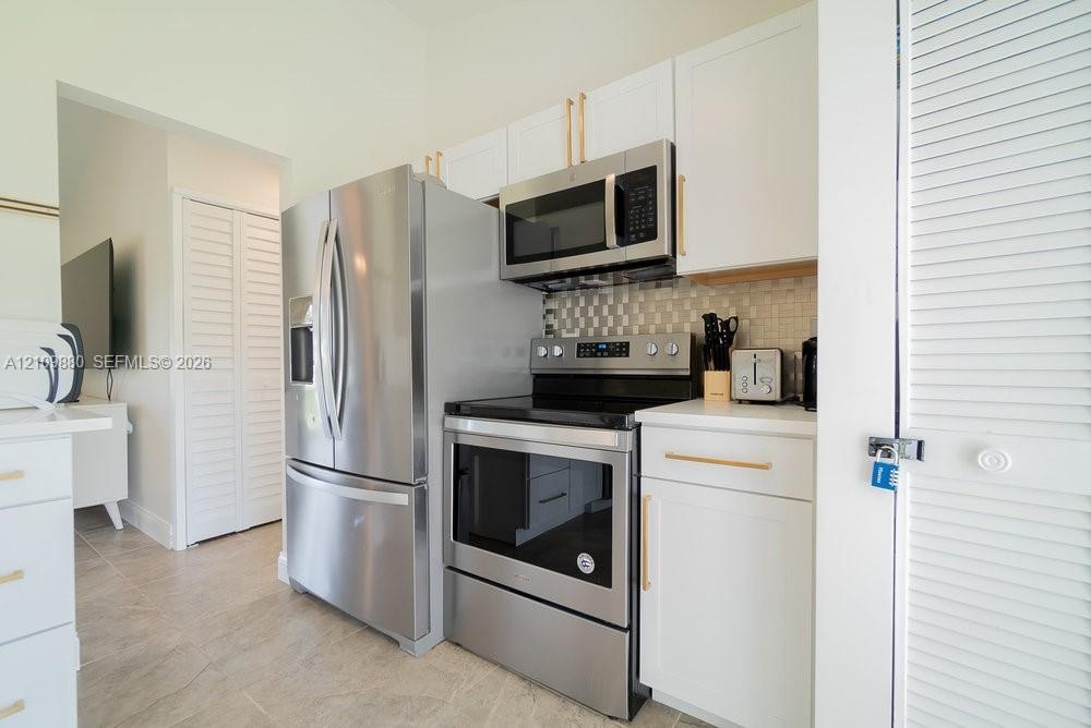 Kitchen featuring white shaker-style cabinetry, stainless steel appliances, brushed gold-tone hardware, a tiled backsplash, and stone-finish counterto