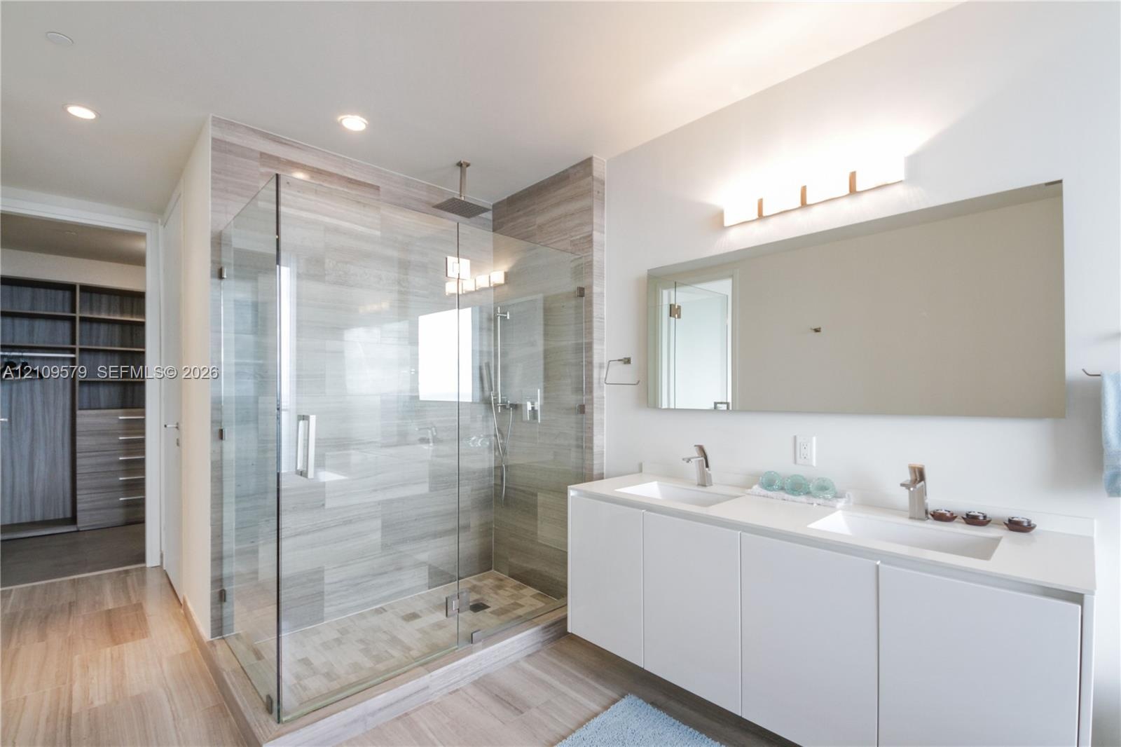 Dual-sink vanity with white cabinetry and rectangular basins, oversized frameless vanity mirror, glass-enclosed walk-in shower with tiled walls and ra