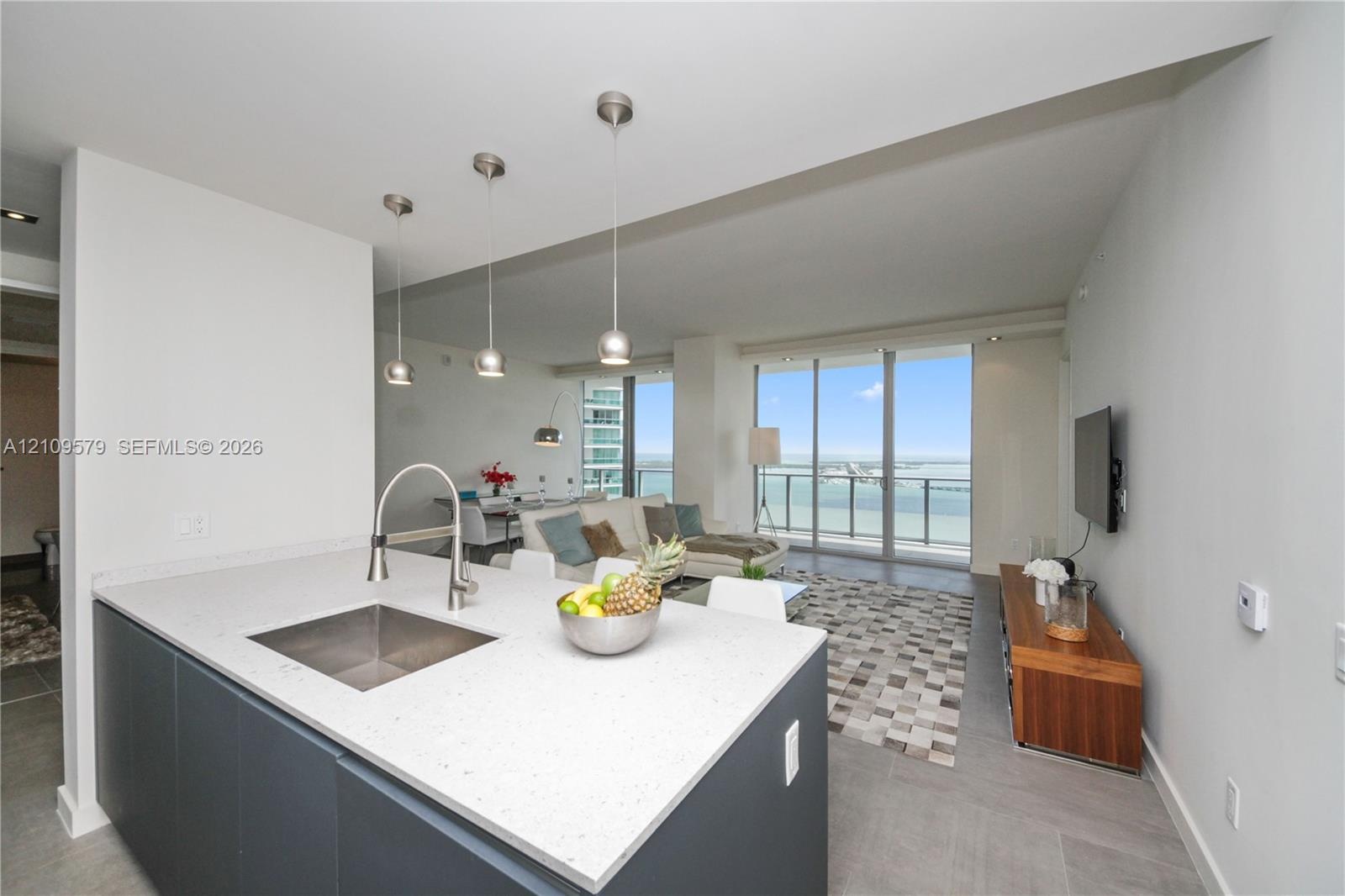 Kitchen island featuring stone countertops, a stainless steel under-mount sink, matte-finish cabinetry, and three pendant light fixtures, with expansi