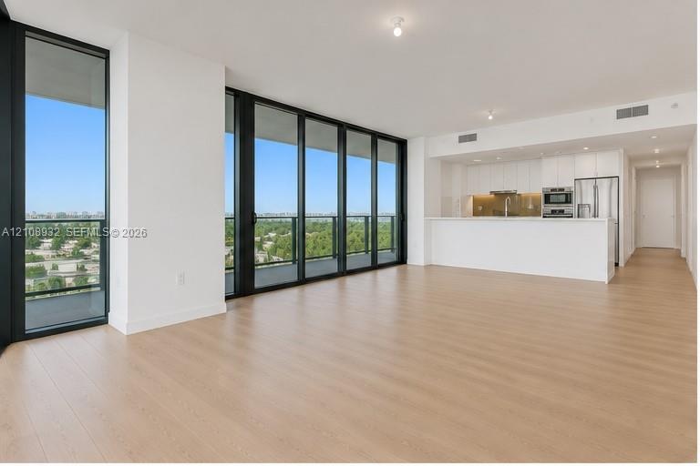 Open-concept living space featuring floor-to-ceiling windows with dark frames, wood-finish flooring, and a kitchen equipped with white flat-panel cabi