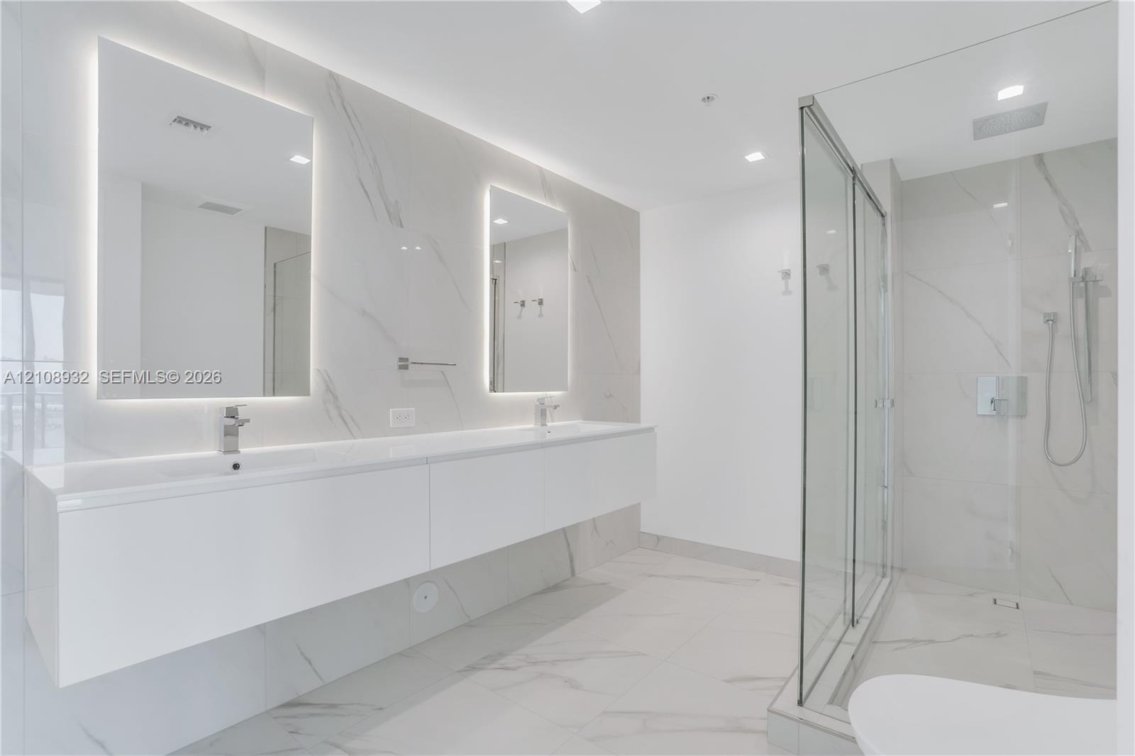 Floating dual vanity with integrated sinks, back-lit mirrors, walk-in glass shower enclosure, and full-height stone-finish wall tiling