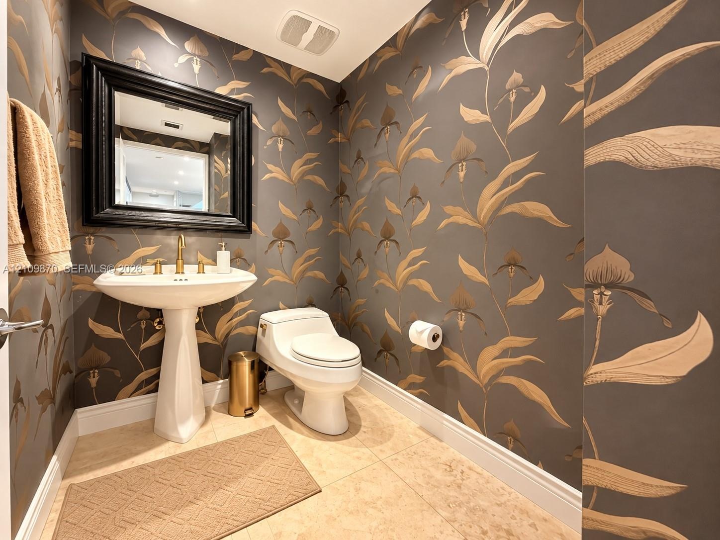 Powder room featuring floral-patterned wallpaper, a white pedestal sink with a brass-finish faucet, a one-piece toilet, and large-format tile flooring