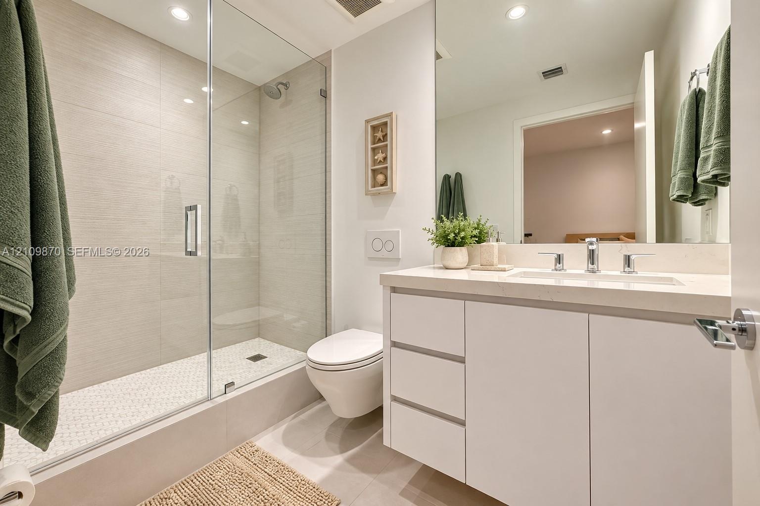 Bathroom featuring a white vanity with an integrated countertop, wall-mounted toilet, walk-in shower with a glass enclosure, and light-toned tile floo