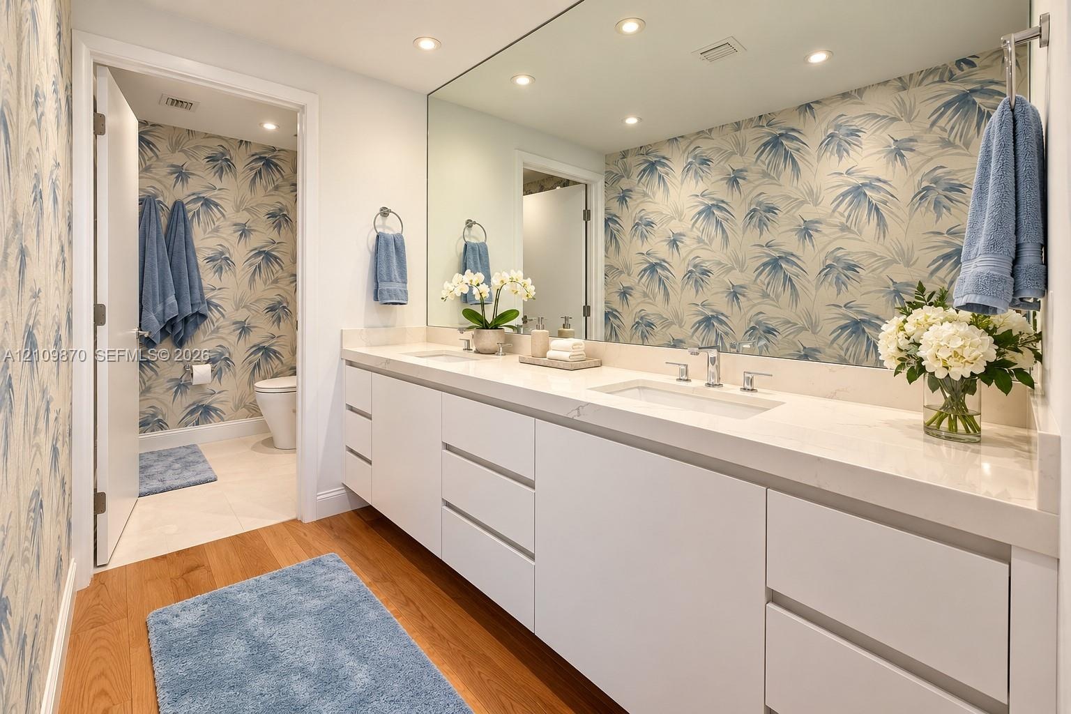 Dual-sink vanity with white cabinetry, light-toned stone countertop, large frameless wall-to-wall mirror, recessed lighting, and wood-finish flooring