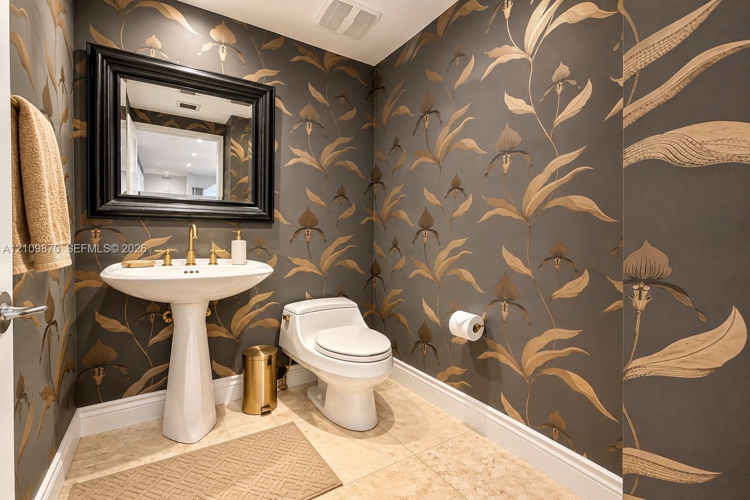 Powder room featuring patterned wallpaper, white pedestal sink, brass-finish widespread faucet, framed wall mirror, and natural stone tile flooring