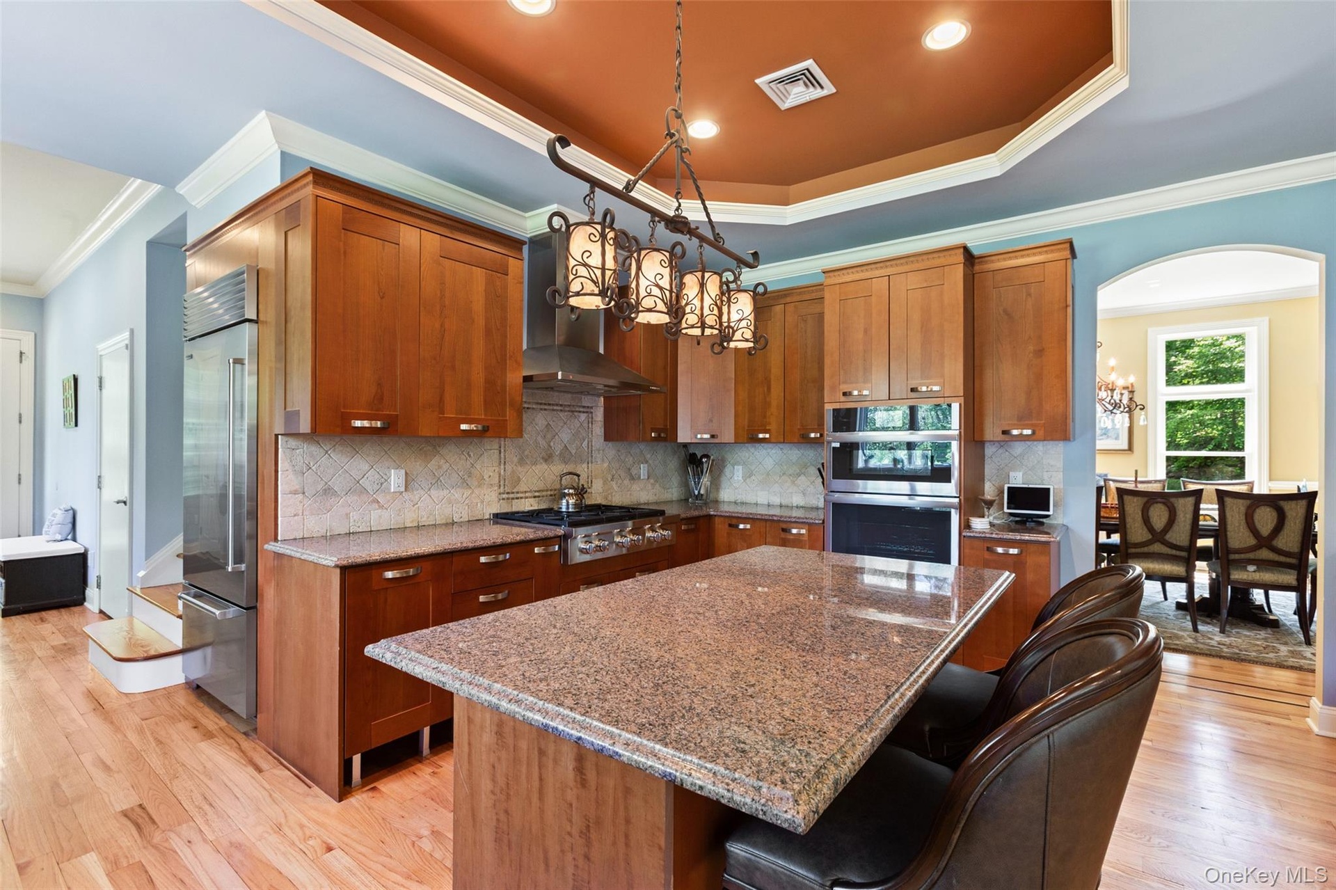 Kitchen with appliances with stainless steel finishes, a chandelier, crown molding, brown cabinetry, and wall chimney range hood