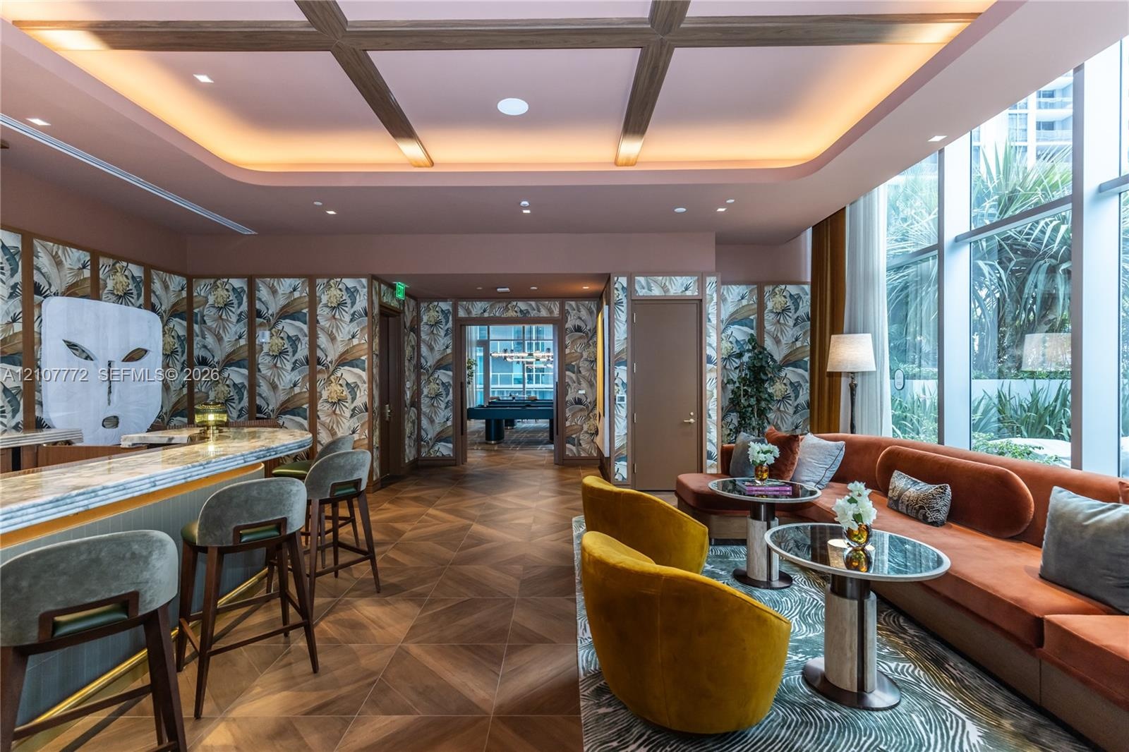 Coffered ceiling with integrated cove lighting, wood-finish flooring in a chevron pattern, floor-to-ceiling windows, built-in bar, and custom wall tre