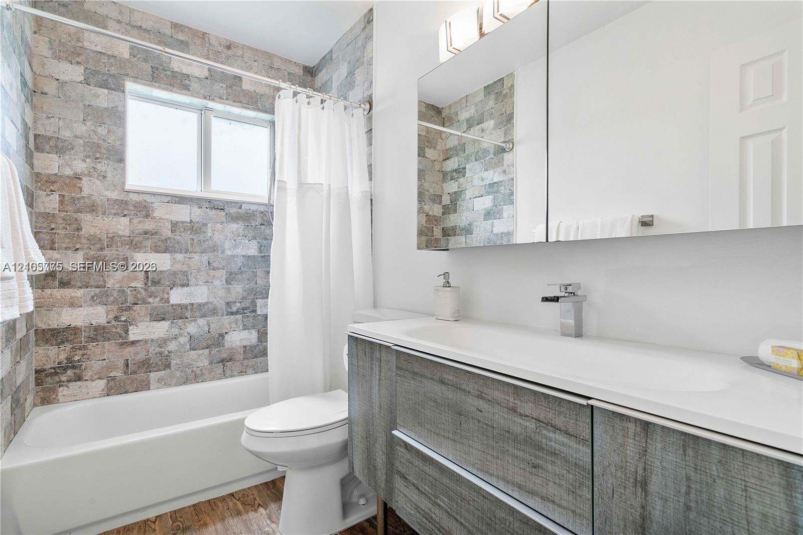 Textured stone tile wall encompassing the bathtub, wood-finish floating vanity, integrated rectangular sink, wall-mounted faucet, and mirrored medicin