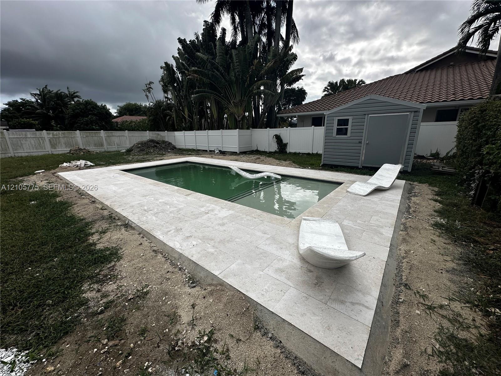 Backyard featuring an in-ground swimming pool with a tanning ledge, light-toned stone-finish paver decking, a white vinyl privacy fence, and a detache